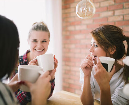 Portrait of joyful young woman enjoying a cup of coffee at home. Smiling pretty girl drinking hot tea in front of the window in winter. Excited woman wearing spectacles and sweater and laughing in an autumn day.