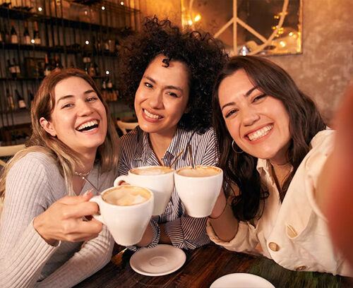 Portrait of joyful young woman enjoying a cup of coffee at home. Smiling pretty girl drinking hot tea in front of the window in winter. Excited woman wearing spectacles and sweater and laughing in an autumn day.