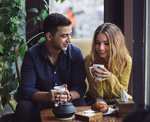 Portrait of joyful young woman enjoying a cup of coffee at home. Smiling pretty girl drinking hot tea in front of the window in winter. Excited woman wearing spectacles and sweater and laughing in an autumn day.
