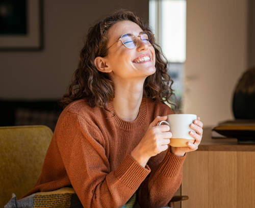 Portrait of joyful young woman enjoying a cup of coffee at home. Smiling pretty girl drinking hot tea in front of the window in winter. Excited woman wearing spectacles and sweater and laughing in an autumn day.